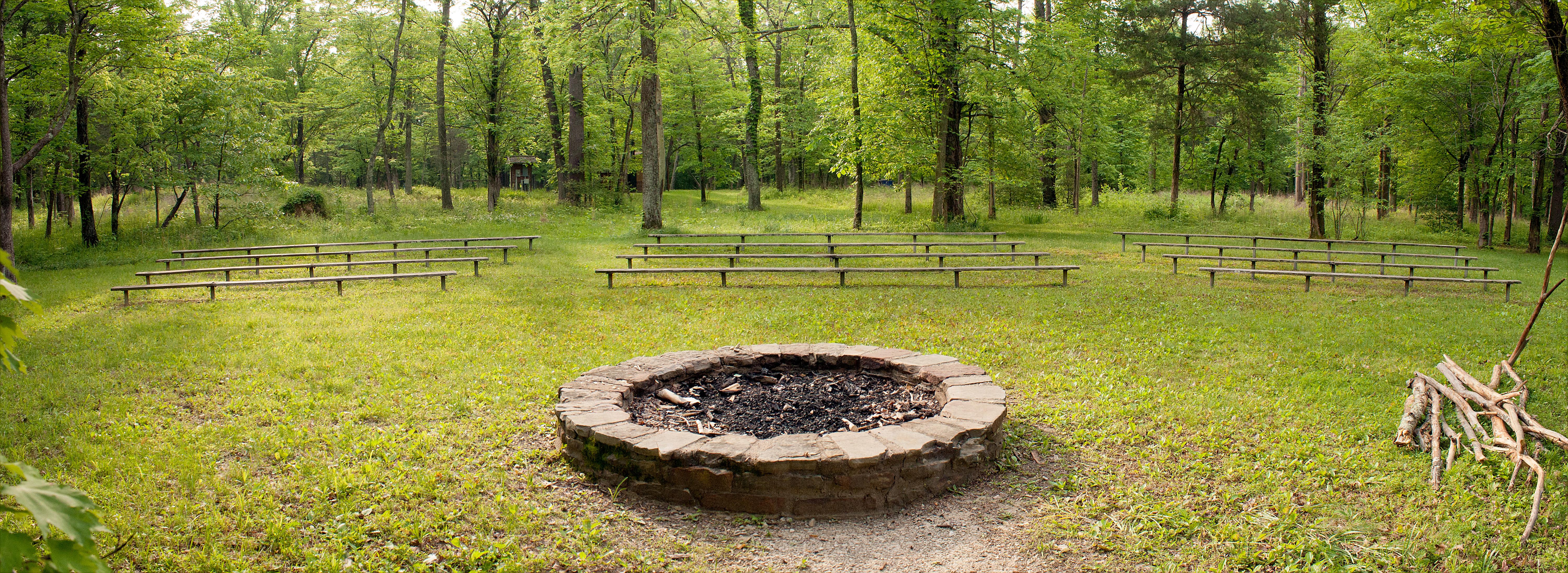 A large stone campfire circle is surrounded by a semicircle of benches in the woods.