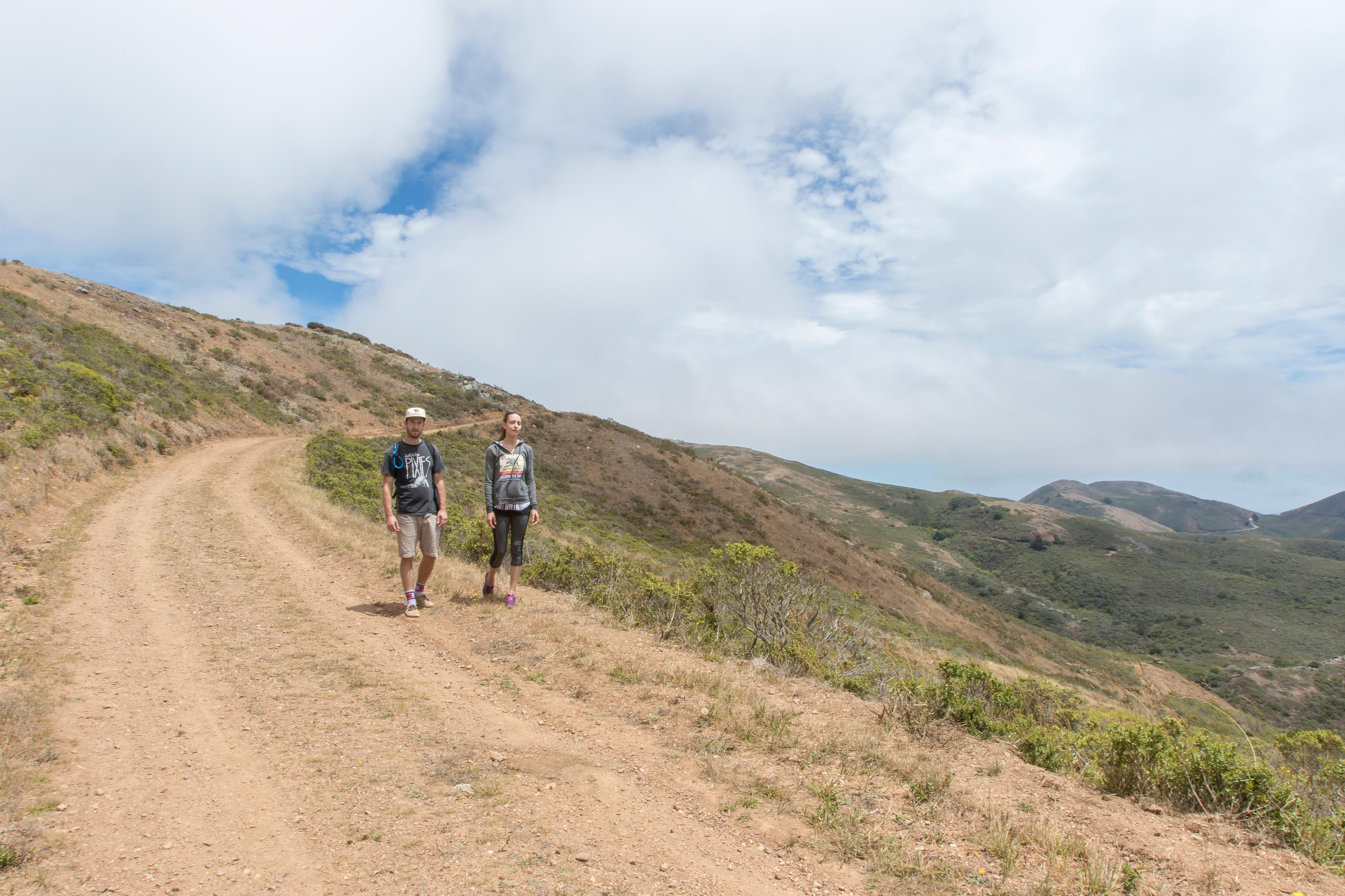 Two hikers walk the ridge along the exposed fire road to Hawk Camp under a cloudy, blue sky.
