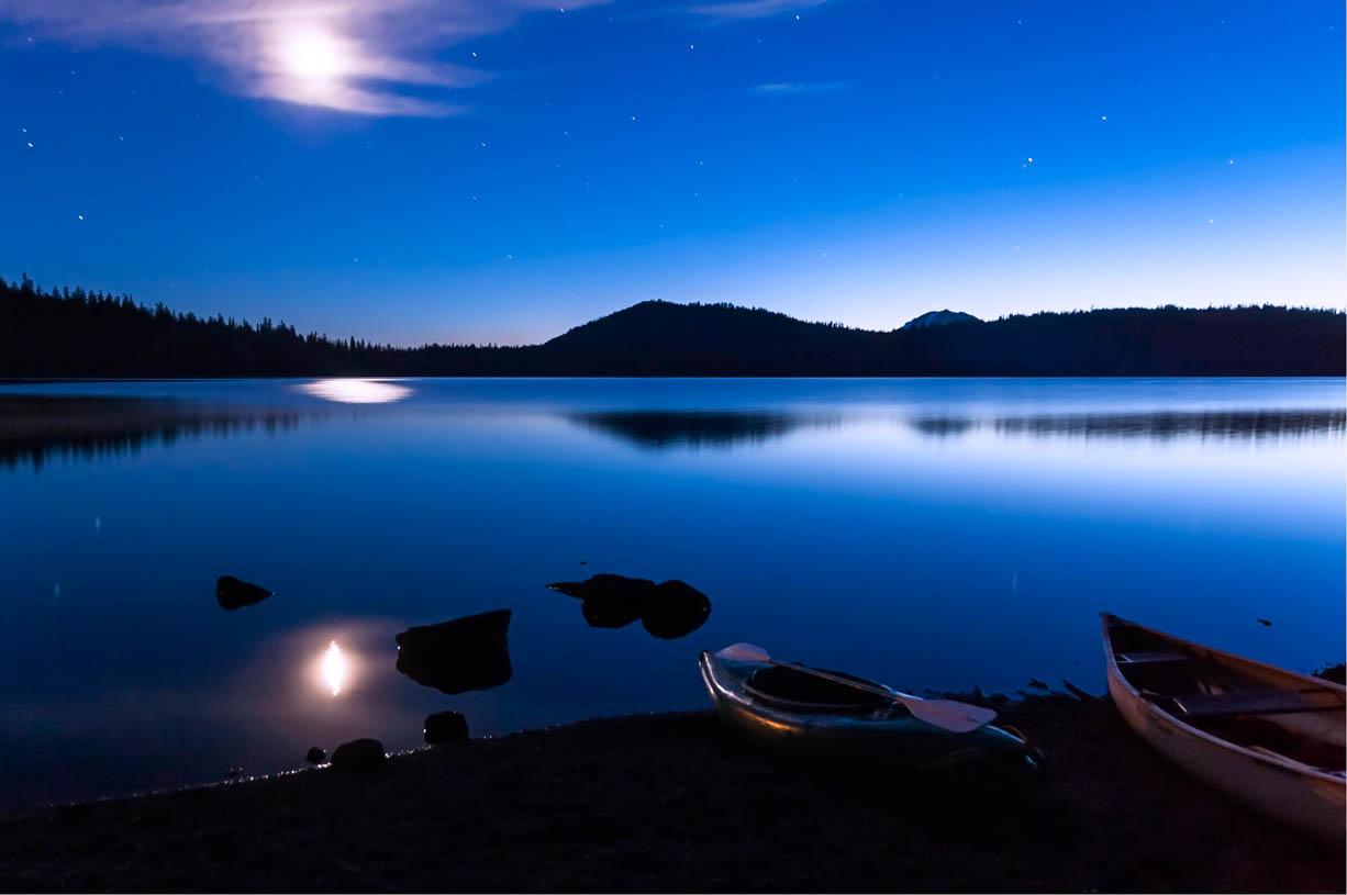 A photo of a kayak and row boat on the short of a moonlit, mountain lake.