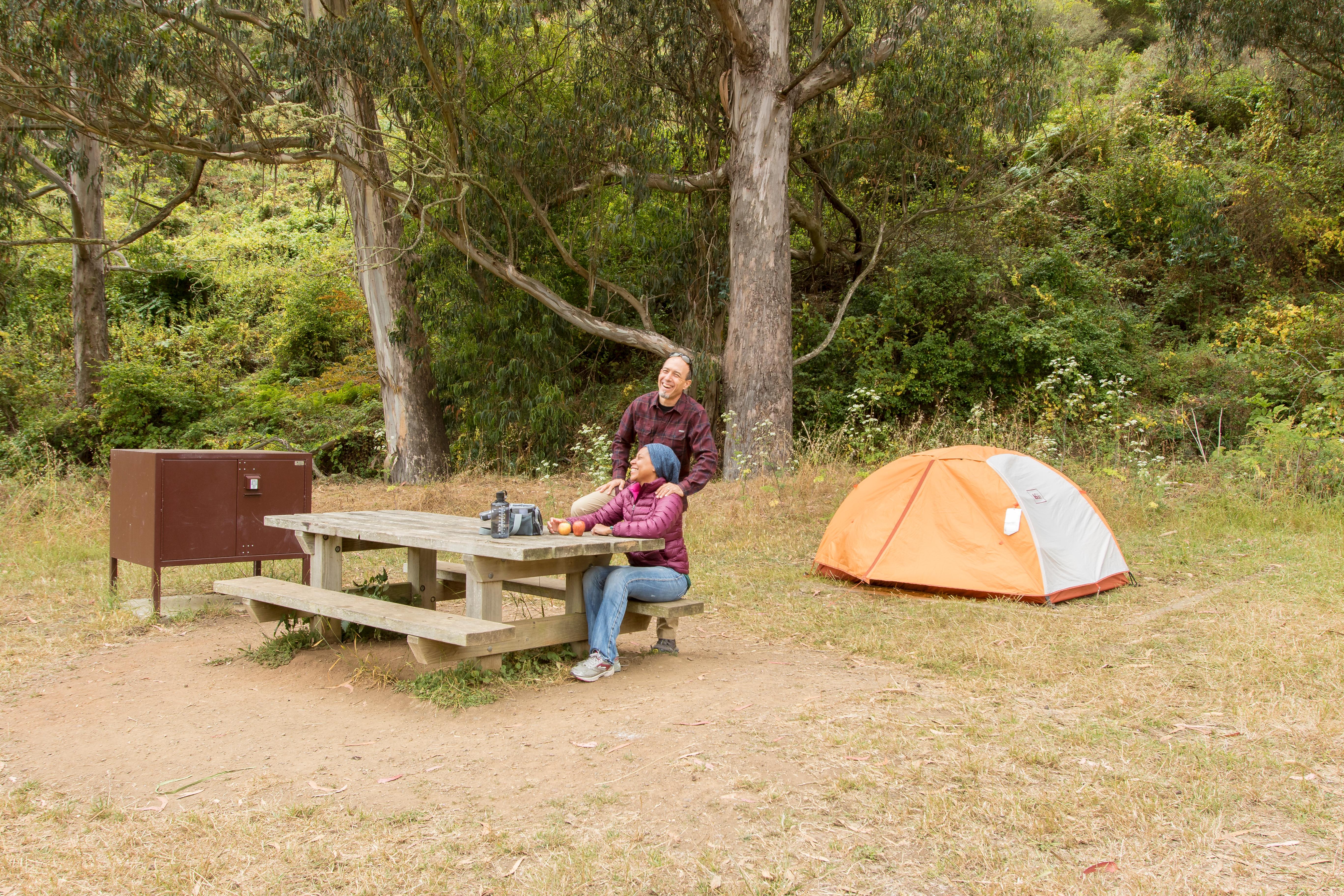 Campers sit at a picnic table next to a food storage box with their tent behind them.