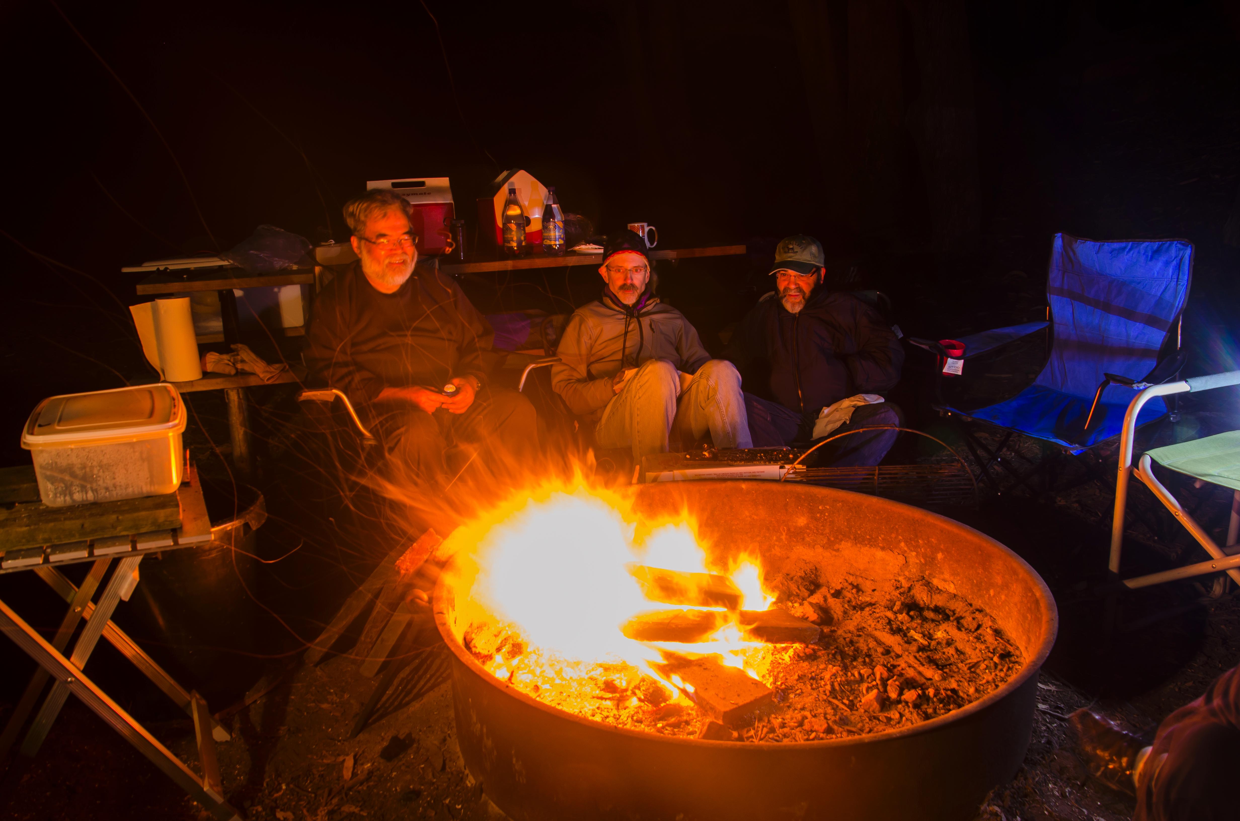 A group of campers sit around the orange glow of a campfire in a fire pit.