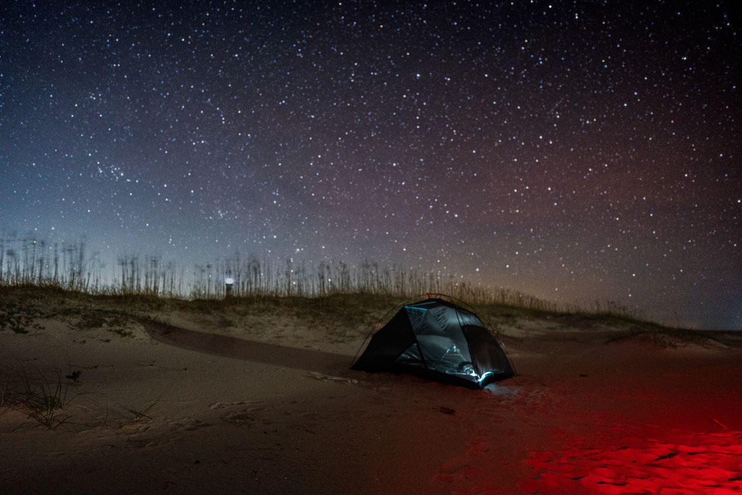 A red light illuminates a tent on the sand, with a star filled sky behind it.