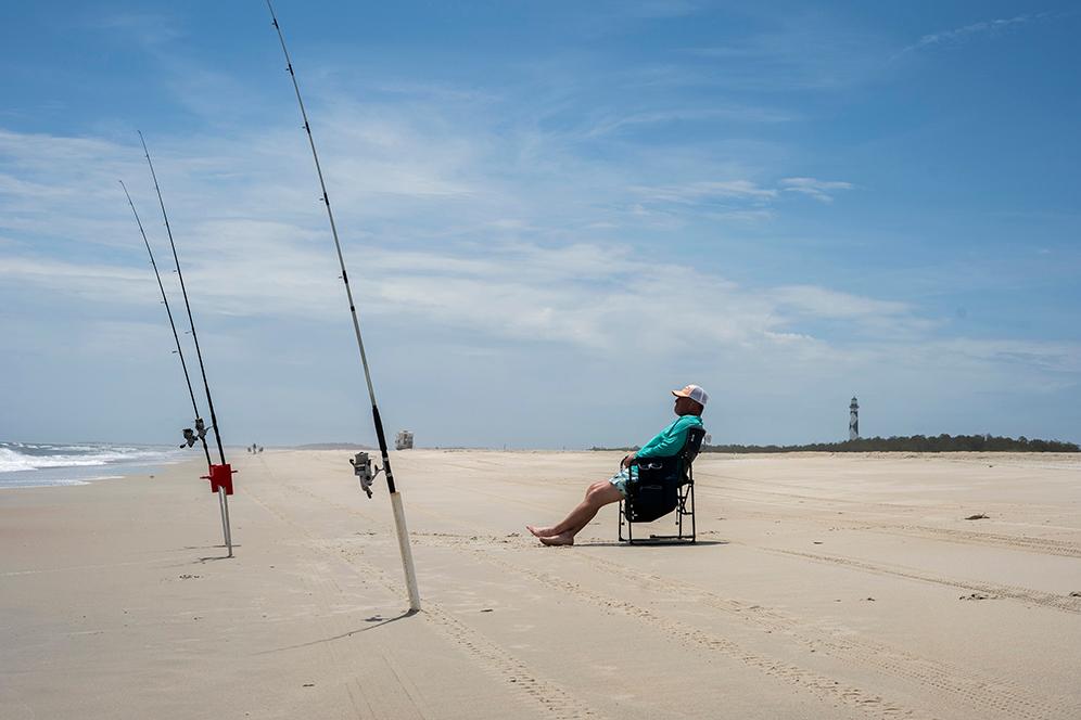 A angler sits in a chair, with two fishing poles in the sand. Lighthouse in background.