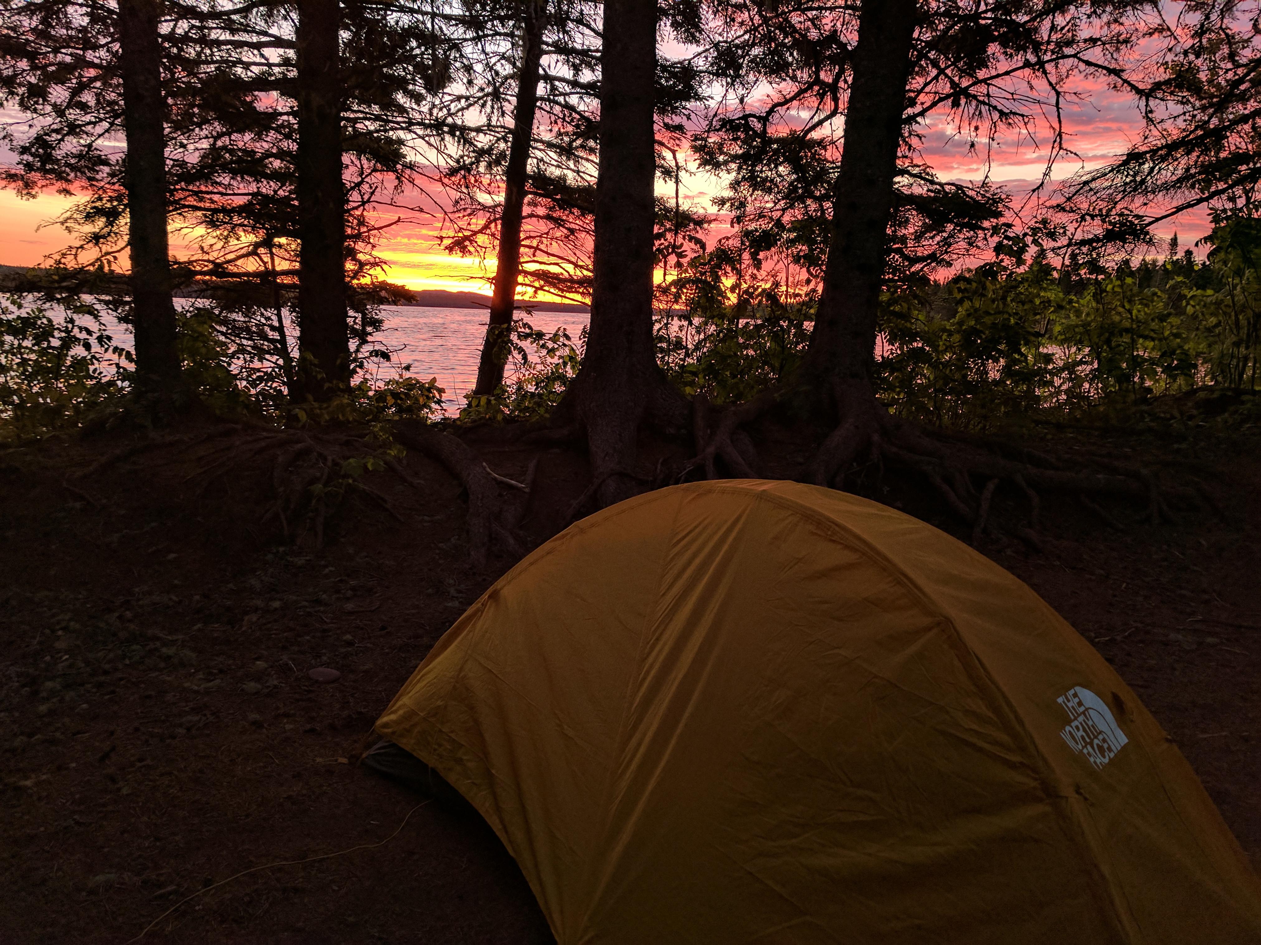 The sun rises through trees on the edge of Feltman Lake. A yellow tent sits in the foreground.