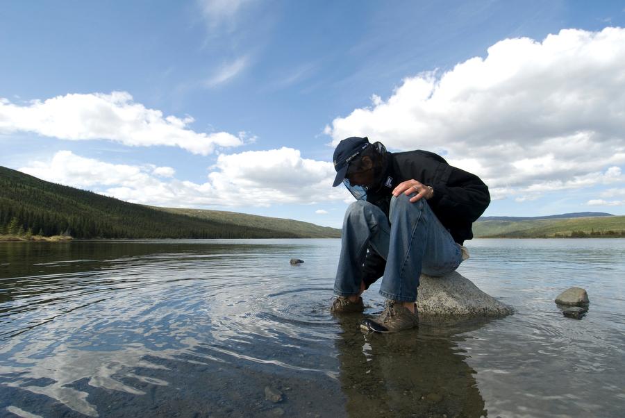 a man sitting on a rock in a lake wearing long pants, a jacket and a head net