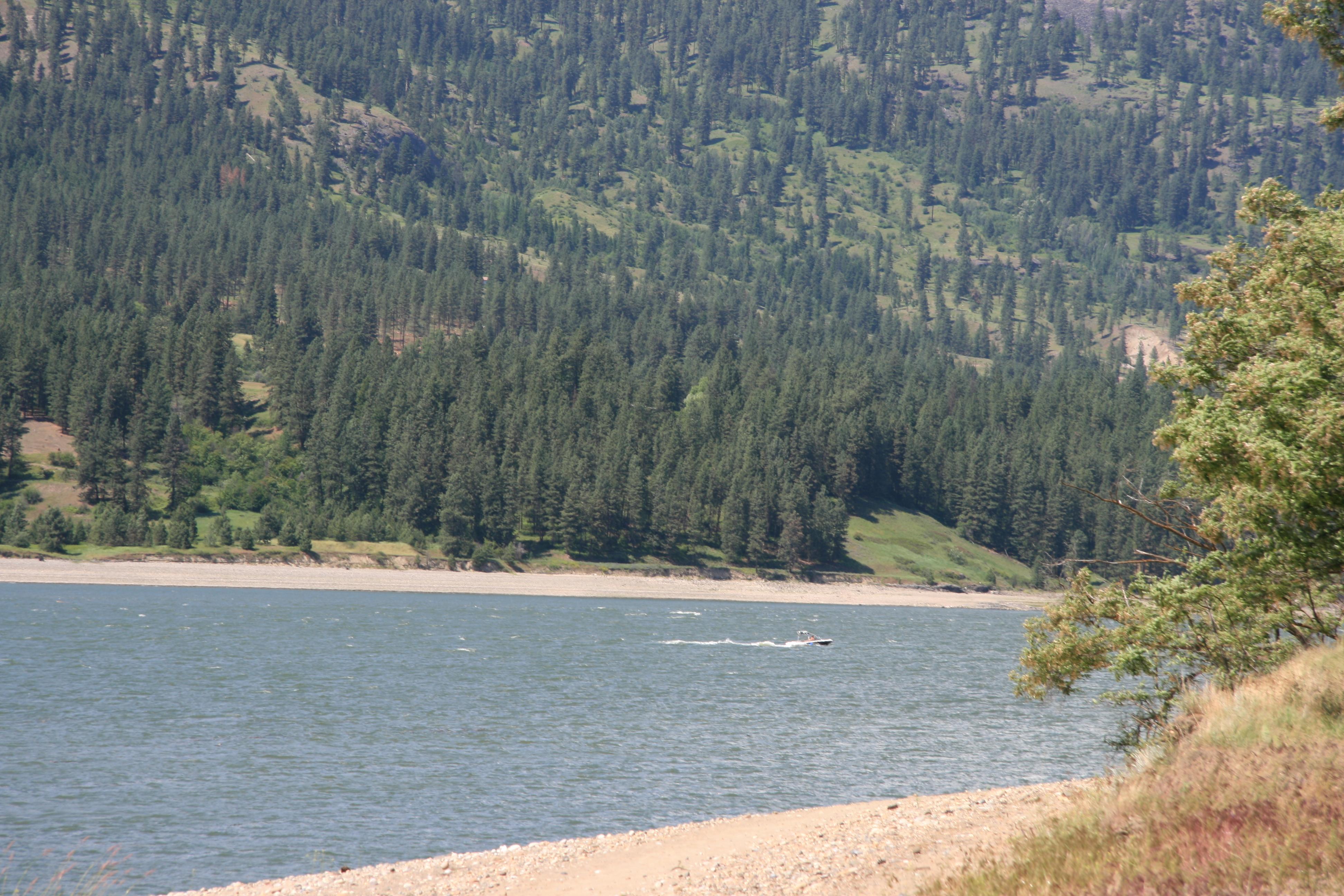 A view of the lake with a boat speeding past, heavy pines on the hillslopes and sand in the fore.