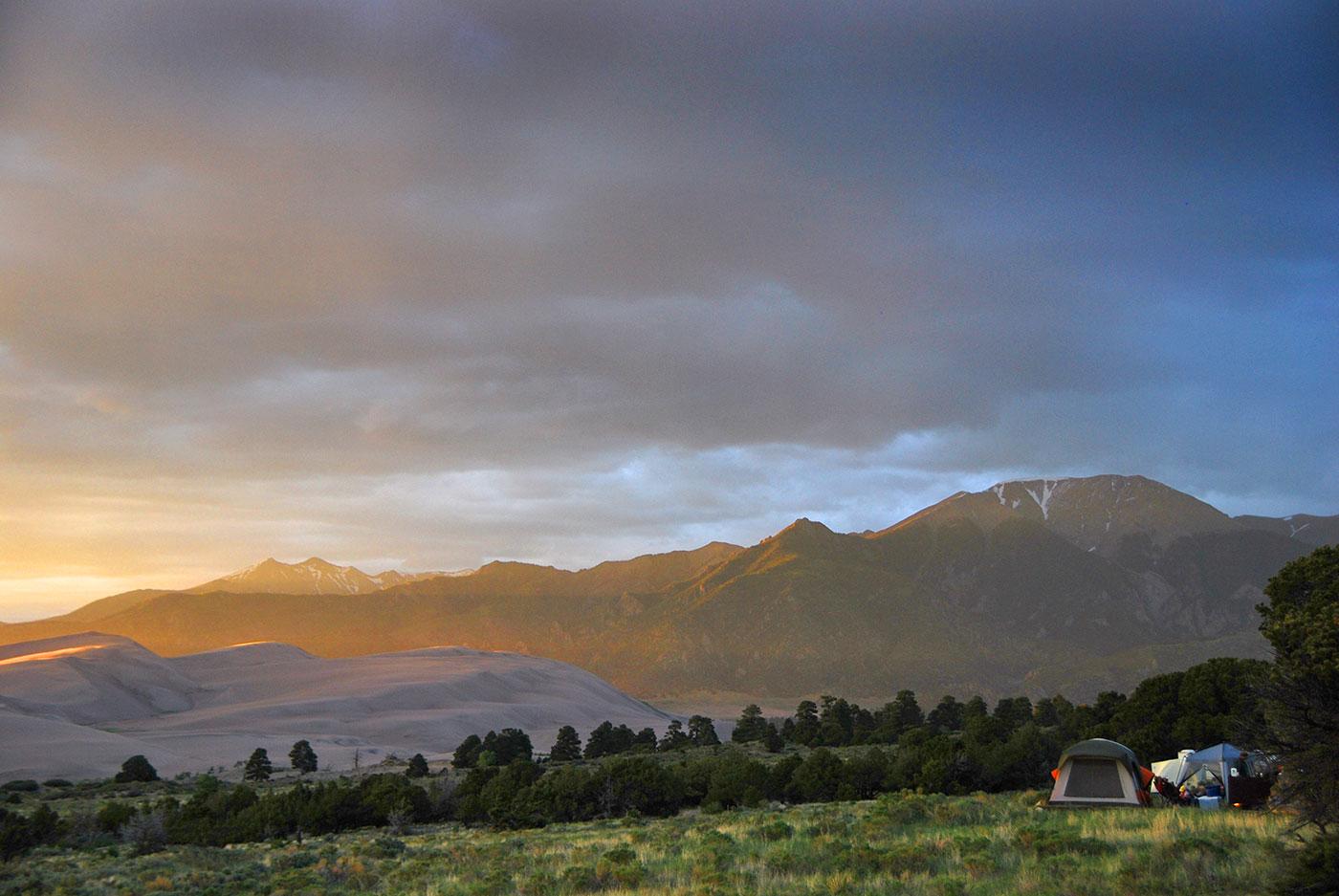 A campsite and tent sit in a scene of grasslands, forest, dunes, and a snow-capped mountain