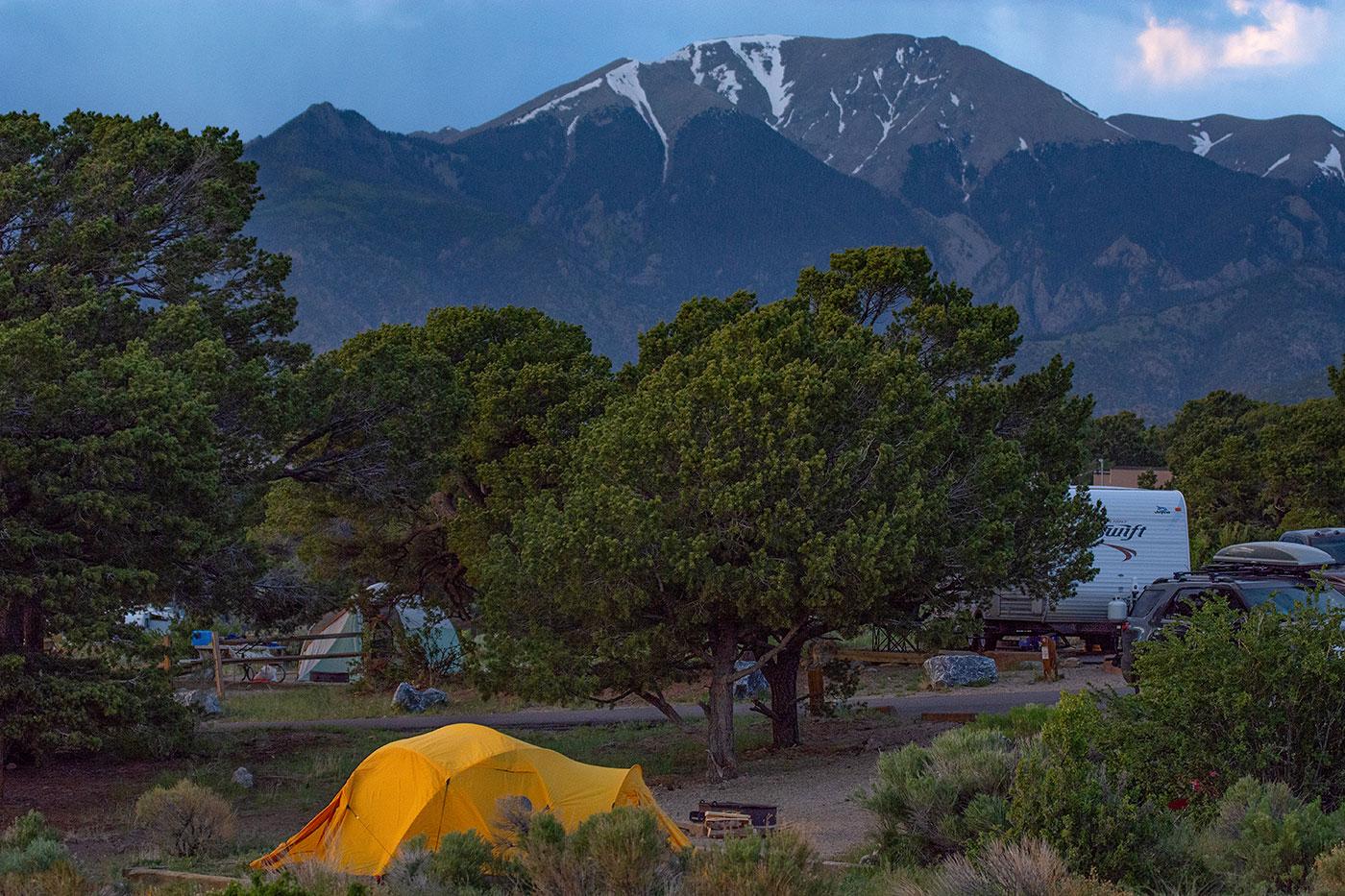 A tent and RV in a campground with small trees, with a snow-capped mountain in background