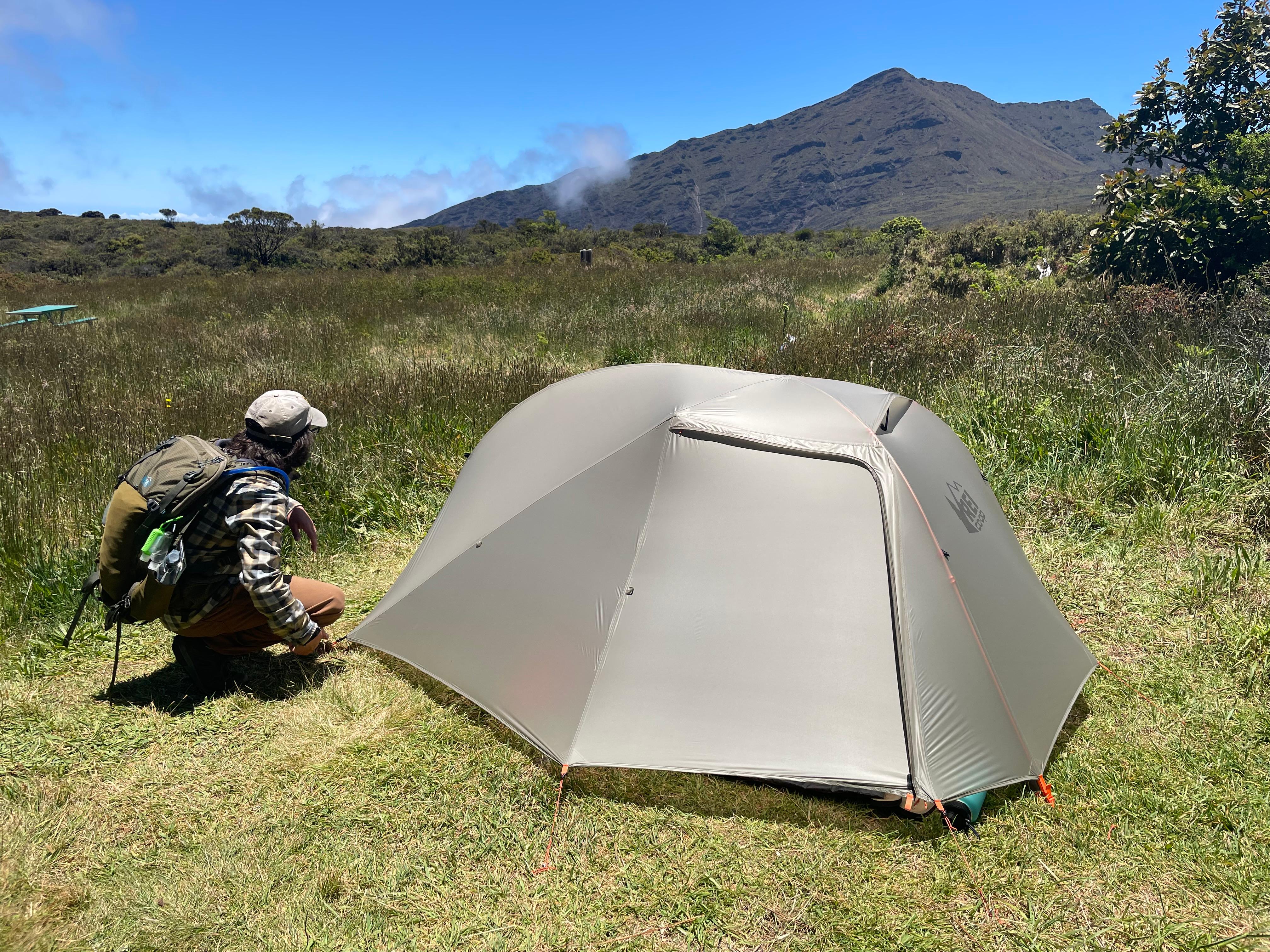 camper setting up tent in grassy area with mountain peak in background